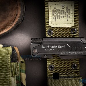 May include: A black multi-tool knife with the engraved text "Best Brother Ever!" and "12.25.2019". The knife is lying on a green belt with a label that reads "WAIST UNDER 30 INCHES". A brown boot and a camouflage pouch are also visible.