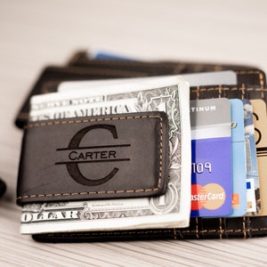 May include: A brown leather money clip with the name "CARTER" engraved in a circle. The money clip is holding a stack of US dollar bills and credit cards.