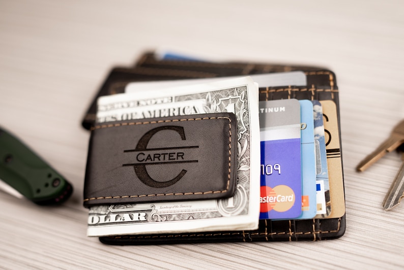 May include: A brown leather money clip with the name "CARTER" engraved in a circle. The money clip is holding a stack of US dollar bills and credit cards.