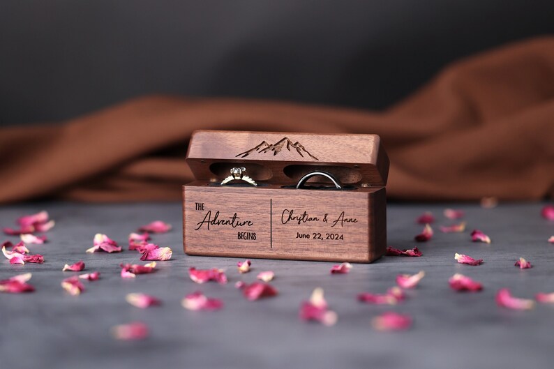 May include: A wooden ring box with a mountain design on the lid. The box is open and has two rings inside. The box is engraved with the text "The Adventure Begins" and "Christian & Anne June 22, 2024".