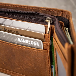 May include: A brown leather bi-fold wallet featuring a zippered compartment, multiple card slots, and a clear ID window. The wallet contains several credit cards, including one labeled Bank One and another labeled The Associates, with visible stitching.