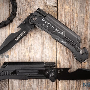 May include: Two black tactical knives with serrated blades and the text "WARTECH." One knife is engraved with "Deputy D. Smith #5237 Stay Safe!" A black paracord lanyard is visible.