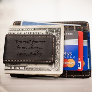 May include: A black leather money clip with a personalized message that reads "You will forever be my always. Love, Ashley". The money clip is holding a dollar bill and several credit cards.