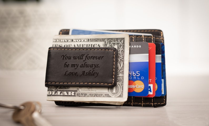 May include: A black leather money clip with a personalized message that reads "You will forever be my always. Love, Ashley". The money clip is holding a dollar bill and several credit cards.
