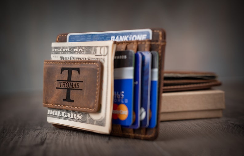 May include: A brown leather wallet with a money clip and a personalized inscription that reads "T Thomas DOLLARS". The wallet is filled with cash and credit cards.