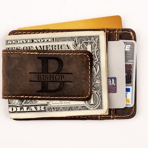 May include: A brown leather wallet with a money clip and a personalized monogram. The monogram is a large "B" with the name "BISHOP" underneath. The wallet is holding a dollar bill and a credit card.