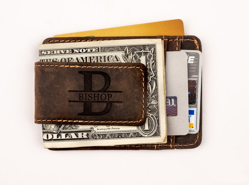 May include: A brown leather wallet with a money clip and a personalized monogram. The monogram is a large "B" with the name "BISHOP" underneath. The wallet is holding a dollar bill and a credit card.