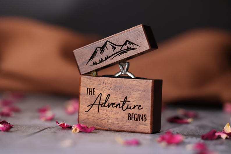 May include: A wooden ring box with a mountain range design on the lid and the text "The Adventure Begins" on the front. The box is open and a silver ring is inside.