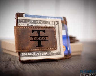 May include: A brown leather money clip wallet featuring a magnetic flap engraved with a large letter T and the name THOMAS centered horizontally. The wallet holds a ten-dollar bill and several credit cards in its slots.