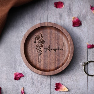 May include: A wooden ring dish with a floral design and the name "Angela" engraved on it. The dish is surrounded by rose petals.