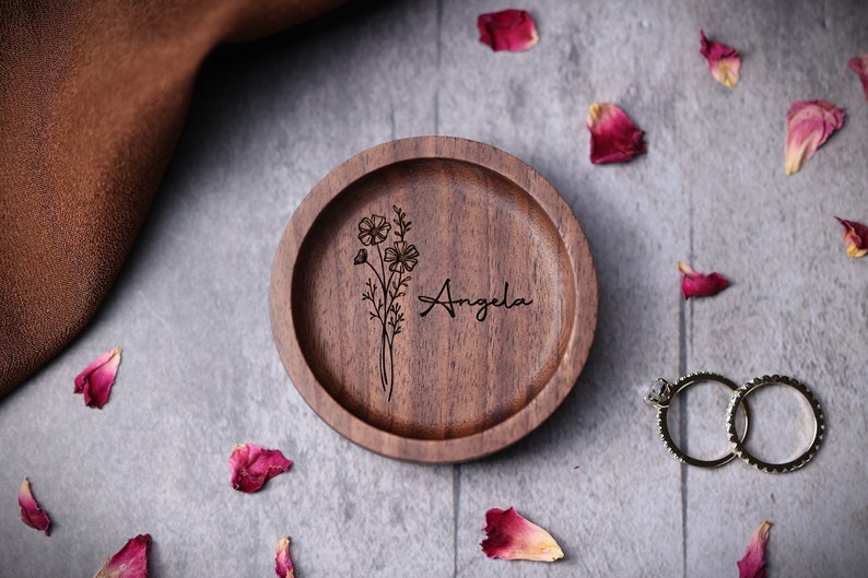 May include: A wooden ring dish with a floral design and the name "Angela" engraved on it. The dish is surrounded by rose petals.