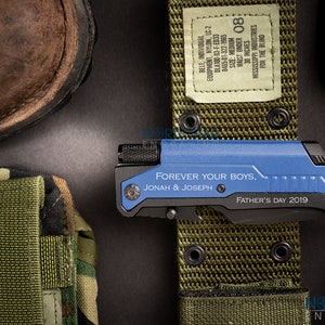 May include: A blue and black multi-tool knife with the inscription "FOREVER YOUR BOYS, JONAH & JOSEPH, FATHER'S DAY 2019." The knife is next to a green belt with a label that reads "WAIST UNDER 30 INCHES." A brown boot and a camouflage pouch are also visible.