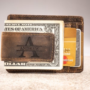 May include: A brown leather wallet with a money clip and a personalized engraved name "ANDREW". The wallet has a worn, distressed look and is holding a dollar bill and credit cards.