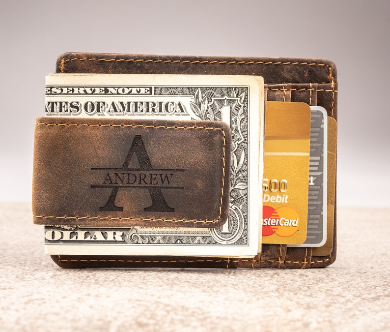May include: A brown leather wallet with a money clip and a personalized engraved name "ANDREW". The wallet has a worn, distressed look and is holding a dollar bill and credit cards.