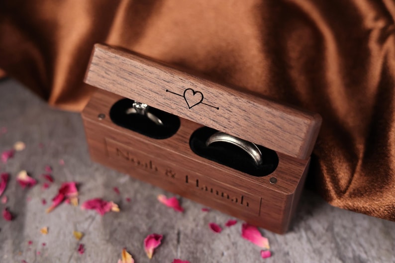 May include: A wooden ring box with two compartments for rings. The box is engraved with the words "Noah & Hannah" and a heart symbol. The box is open and two rings are visible inside.