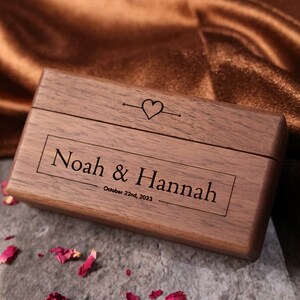 May include: A wooden ring box with the names "Noah & Hannah" and the date "October 22nd, 2023" engraved on the lid. The box is closed and has a heart symbol on the top.