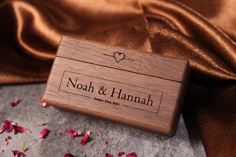 May include: A wooden ring box with the names "Noah & Hannah" and the date "October 22nd, 2023" engraved on the lid. The box is closed and has a heart symbol on the top.