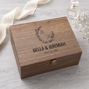 May include: A rectangular dark brown wooden box with a hinged lid and a metal latch. The lid features a laser-engraved floral wreath surrounding the letters B + J, followed by the text BELLA & JEREMIAH and the date JULY 25, 2025.