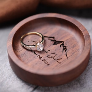 May include: A wooden ring dish with a mountain design and the text "A & Julie October 08, 2024" engraved on it. A gold ring with a diamond is sitting on the dish.