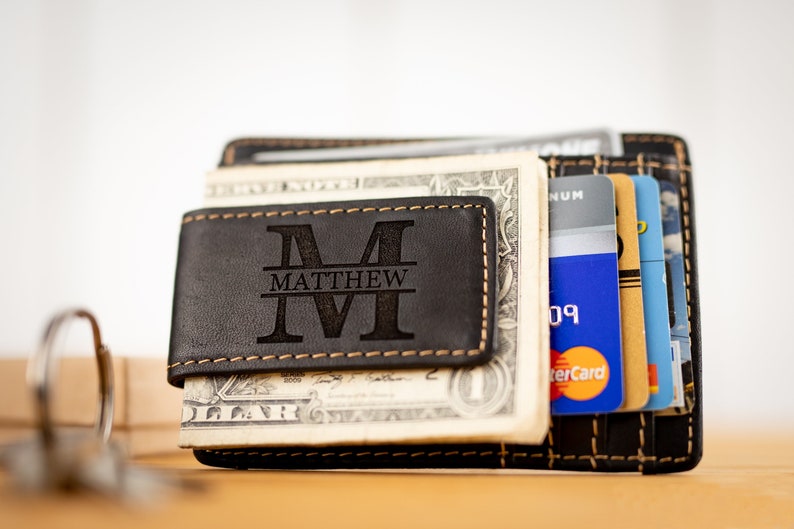 May include: A black leather wallet with a money clip and a personalized monogram. The monogram is a large "M" with the name "MATTHEW" underneath. The wallet is filled with cash and credit cards.