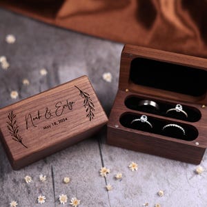 May include: Wooden ring boxes, one open and one closed, with the names "Noah & Evelyn" and the date "May 18, 2024" engraved on the closed box. The open box displays three rings. The boxes are on a gray surface.