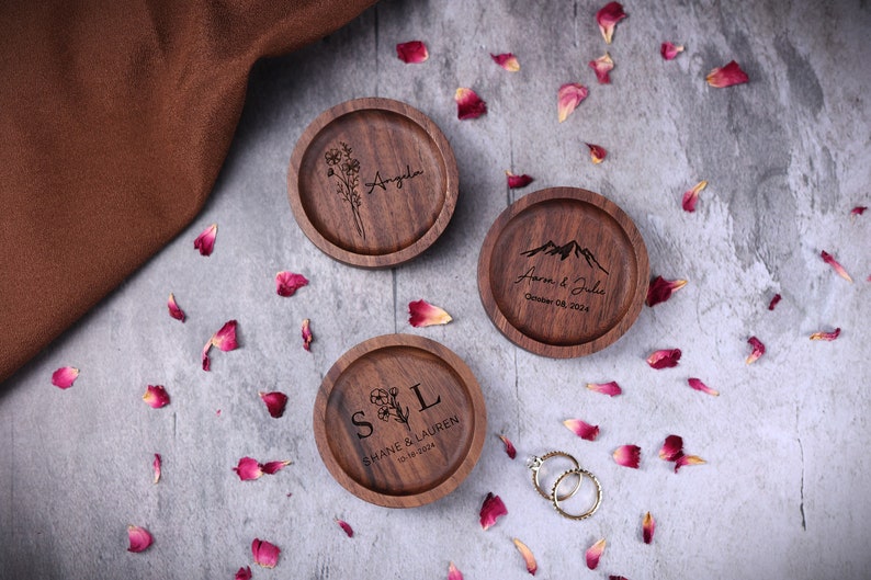 May include: Three wooden ring boxes with engraved designs. The first box has a floral design and the name "Angela" engraved. The second box has a mountain design and the text "Aaron & Julie, October 08, 2024". The third box has a floral design and the text "S&L, Shane & Lauren, 03-18-2024".
