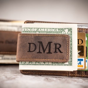 May include: A brown leather money clip with the initials "DMR" engraved on the front. The clip is holding a stack of cash and credit cards.