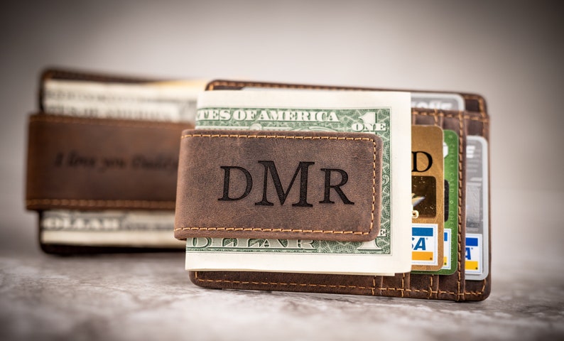 May include: A brown leather money clip with the initials "DMR" engraved on the front. The clip is holding a stack of cash and credit cards.
