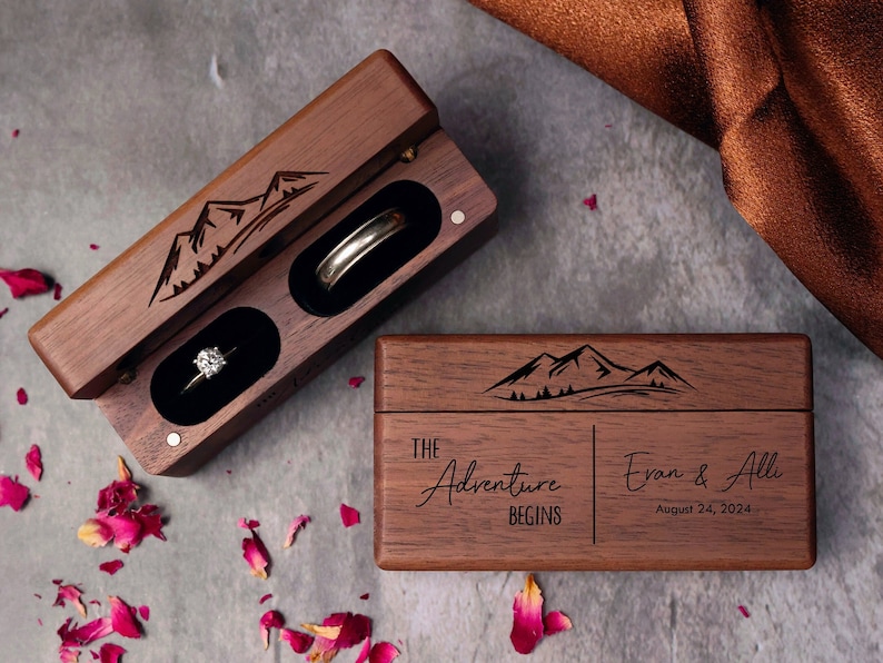 May include: A wooden ring box with a mountain scene engraved on the lid. The box is open and has two compartments, one with a diamond ring and the other with a wedding band. The box also has the text "The Adventure Begins" and "Evan & Alli August 24, 2024" engraved on the front.