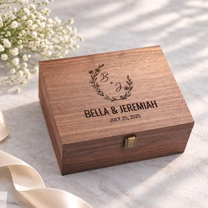 May include: A rectangular dark brown wooden keepsake box with a metal latch. The lid features a laser-engraved floral wreath surrounding the initials B + J, followed by the text BELLA & JEREMIAH and the date JULY 25, 2025.