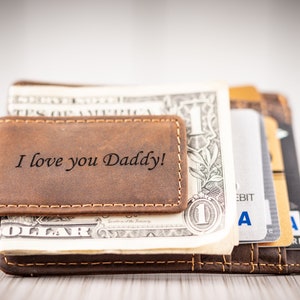 May include: A brown leather money clip with the inscription "I love you Daddy!"  The clip is holding a stack of US dollar bills and credit cards.