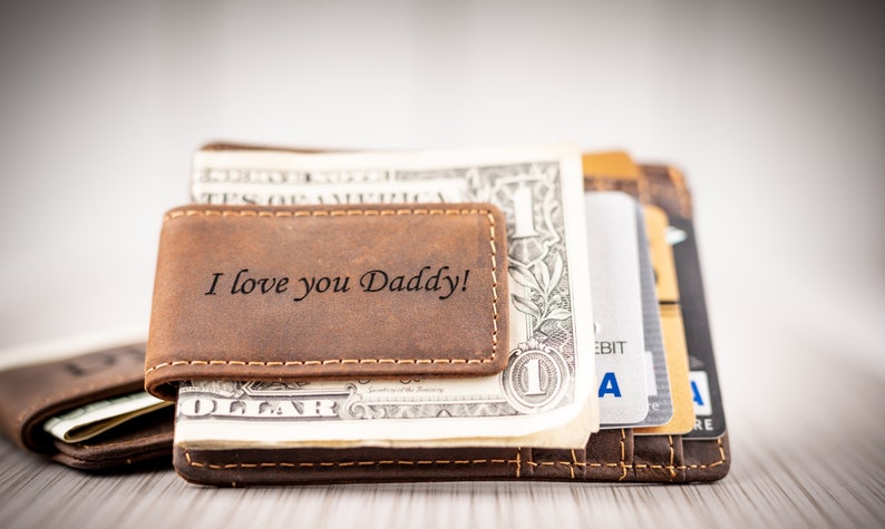 May include: A brown leather money clip with the inscription "I love you Daddy!"  The clip is holding a stack of US dollar bills and credit cards.