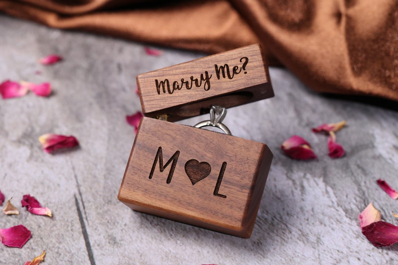 May include: A wooden ring box with the words "Marry Me?" engraved on the lid and the initials "M&L" engraved on the front. The box is open and a silver ring is inside.