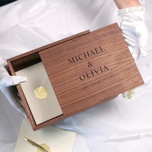 May include: A wooden box with a sliding lid, engraved with the names "MICHAEL & OLIVIA" and the date "09-27-2025" and location. The box is open, revealing a cream-colored envelope with a wax seal. The box is held by gloved hands.