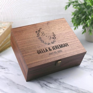 May include: A wooden box with a floral wreath design and the initials "B" and "J". The box is engraved with the names "BELLA & JEREMIAH" and the date "JULY 25, 2025". The box has a brass clasp.