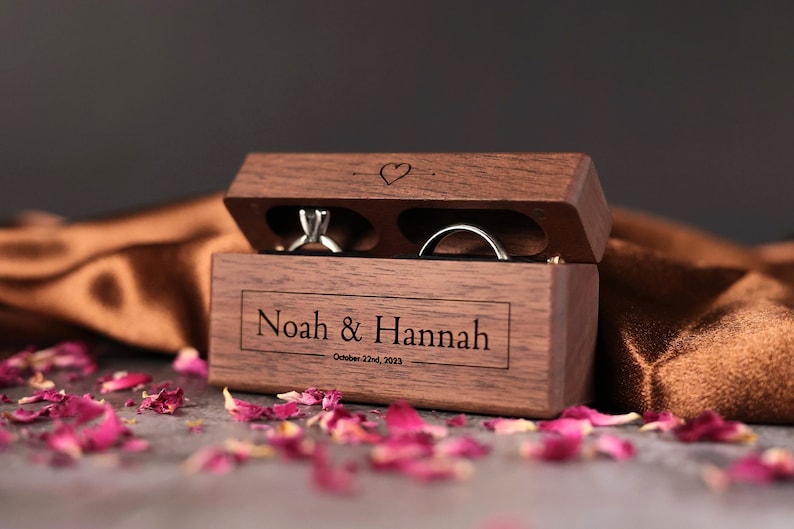May include: A wooden ring box with two compartments for rings. The box is engraved with the names "Noah & Hannah" and the date "October 22nd, 2023".
