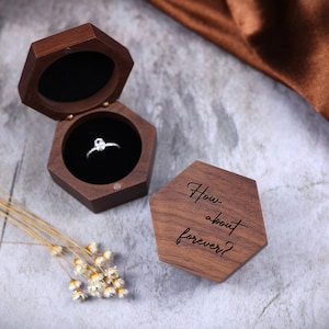 May include: A hexagonal wooden ring box. The box is open and a diamond ring is inside. The box is engraved with the words "How about forever?"