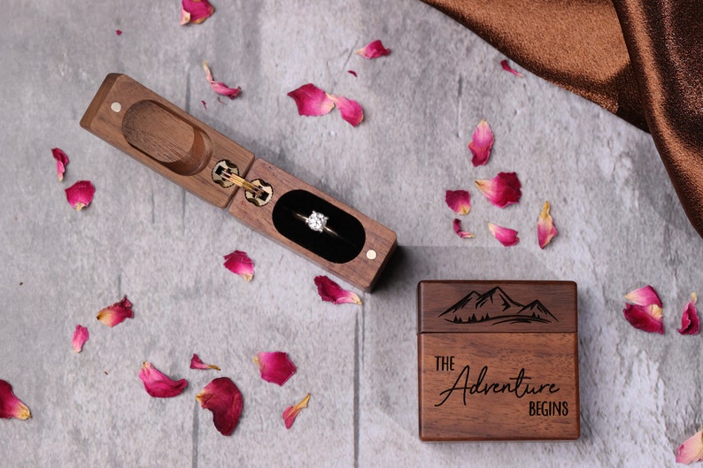 May include: A wooden ring box with a sliding lid. The box is engraved with the words "The Adventure Begins" and a mountain range. The box is open and a diamond ring is inside.