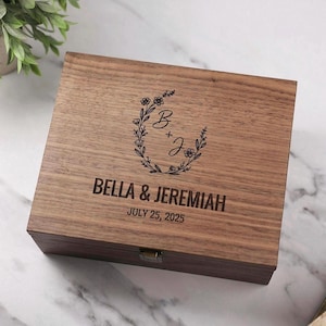 May include: A wooden box with a floral wreath design and the initials "B + J" engraved on the lid. The box is personalized with the names "BELLA & JEREMIAH" and the date "JULY 25, 2025". The box has a rectangular shape and a metal clasp.