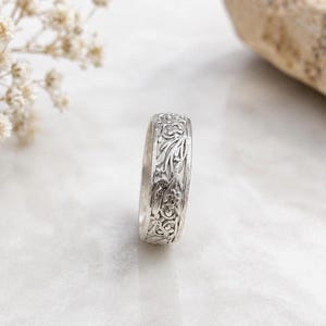May include: A silver ring featuring an intricate, embossed floral and vine pattern covering the entire outer surface. The band has a polished metallic finish with detailed engravings of small flowers and winding leaves throughout the design.