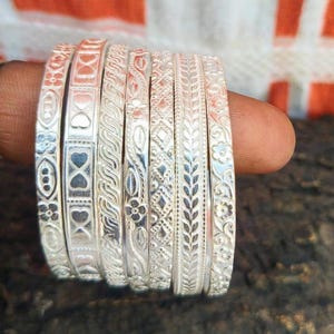 Op de afbeelding: Een verzameling zilveren armbanden met ingewikkelde reliëfdesigns. Elke armband heeft een uniek patroon, waaronder bloemen-, geometrische en bladmotieven. De armbanden worden op een vinger getoond, wat hun gedetailleerde vakmanschap en reflecterende oppervlak benadrukt.