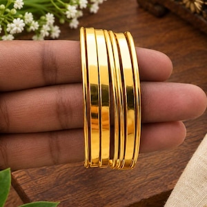 May include: A set of four polished gold-tone bangles with a smooth, reflective surface and a rounded profile. The bracelets are stacked together, showcasing a uniform metallic finish and a simple, clean design without any additional embellishments.