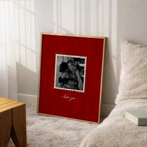 K&ouml;nnte beinhalten: Ein gerahmter Druck mit rotem Hintergrund und einer Schwarz-Wei&szlig;-Fotografie von zwei sich umarmenden Personen. Das Foto befindet sich in einem postkarten&auml;hnlichen Rahmen, darunter die Worte "i love you" in Kursivschrift. Der Rahmen ist goldfarben.