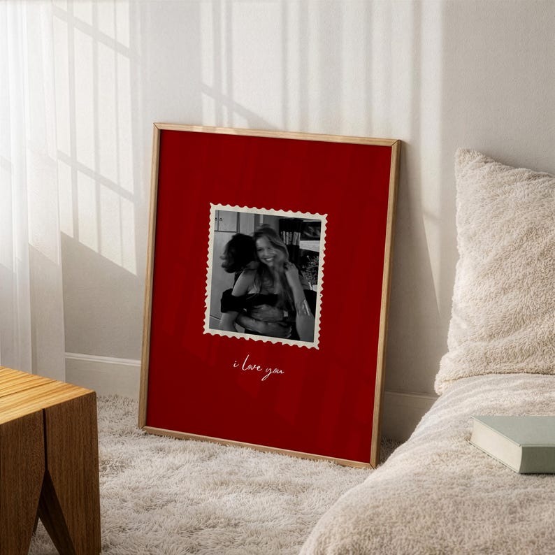 K&ouml;nnte beinhalten: Ein gerahmter Druck mit rotem Hintergrund und einer Schwarz-Wei&szlig;-Fotografie von zwei sich umarmenden Personen. Das Foto befindet sich in einem postkarten&auml;hnlichen Rahmen, darunter die Worte "i love you" in Kursivschrift. Der Rahmen ist goldfarben.