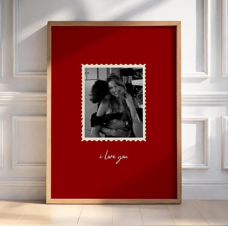 K&ouml;nnte beinhalten: Ein gerahmter Druck mit einem leuchtend roten Hintergrund, der ein Schwarz-Wei&szlig;-Foto von zwei sich umarmenden Personen zeigt, das in einem Rahmen im Postkartenstil eingefasst ist. Die Worte "i love you" stehen darunter in Schreibschrift.