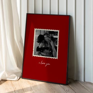 K&ouml;nnte beinhalten: Ein gerahmter Druck mit schwarzem Rahmen. Der Druck hat einen roten Hintergrund mit einem Schwarz-Wei&szlig;-Foto im Poststempelstil. Die Worte "i love you" sind darunter in Kursivschrift geschrieben.