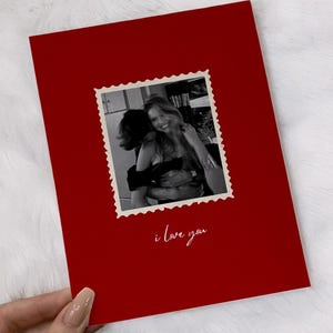 K&ouml;nnte beinhalten: Eine rote Gru&szlig;karte mit einem Schwarz-Wei&szlig;-Foto in einem Briefmarkenrahmen. Das Foto zeigt zwei Personen, die sich umarmen. Die Worte "i love you" sind in Kursivschrift am unteren Rand der Karte geschrieben.