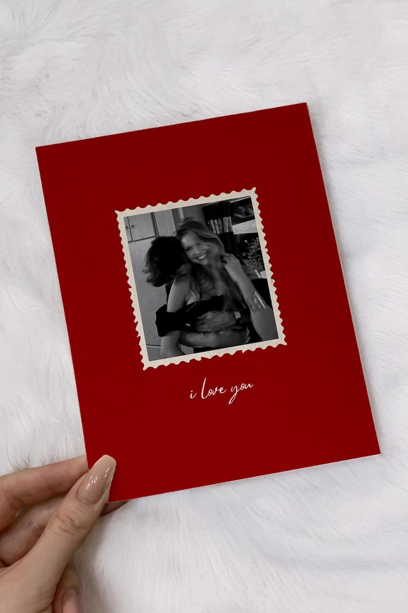 K&ouml;nnte beinhalten: Eine rote Gru&szlig;karte mit einem Schwarz-Wei&szlig;-Foto in einem Briefmarkenrahmen. Das Foto zeigt zwei Personen, die sich umarmen. Die Worte "i love you" sind in Kursivschrift am unteren Rand der Karte geschrieben.