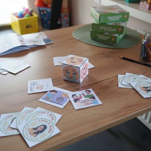 May include: A wooden table displays a variety of educational materials. A cube with emoji faces sits near illustrated cards, stickers, and a stack of green boxes. A jar of colored pencils and an open book are also present.