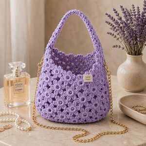 Könnte beinhalten: Eine hellviolette Häkeltasche mit einem sich wiederholenden Gänseblümchenmuster, einem einzelnen Tragegriff und einem goldfarbenen Metallketten-Schulterriemen. Ein kleines rechteckiges Stoffetikett mit dem Text hand made ist vorne angebracht.